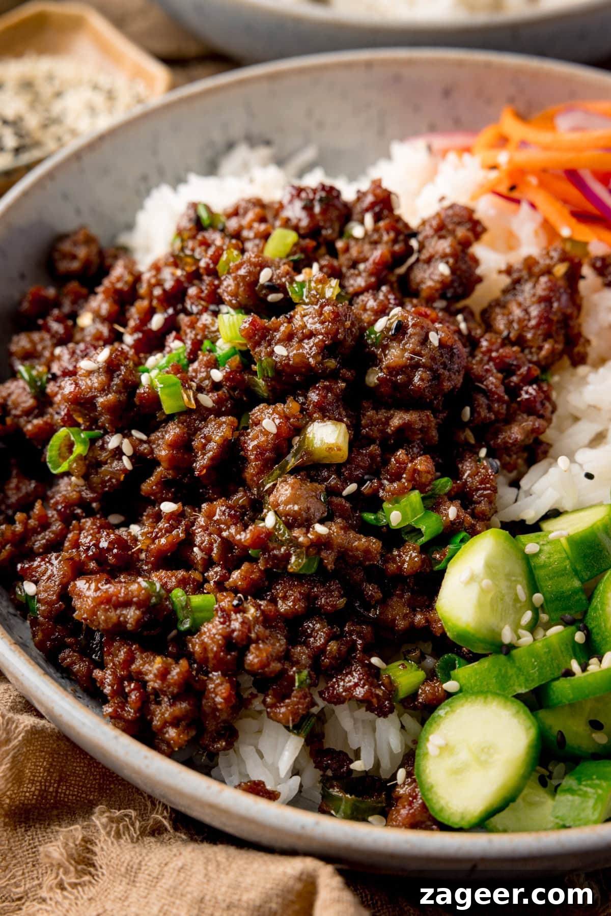 Very close up side-on photo of a light blue speckled bowl filled with Vietnamese-style minced beef on a bed of rice, with pickled strips of red onion and carrot. There are also chunks of baby cucumber and a sprinkling of black and white sesame seeds in the dish.
