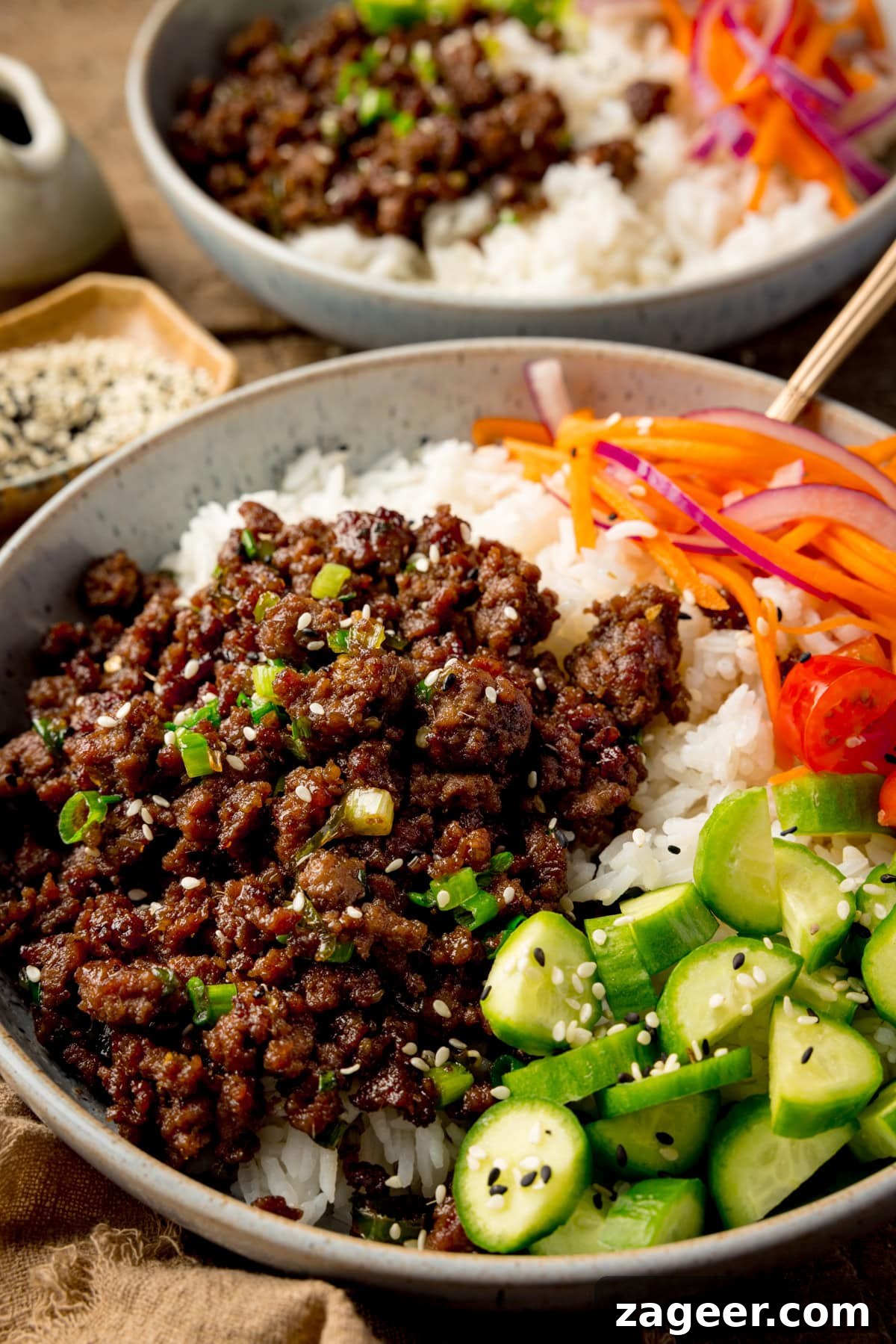 Close up side-on photo of a light blue speckled bowl filled with Vietnamese-style minced beef on a bed of rice, with pickled strips of red onion and carrot. There are also chunks of baby cucumber and chopped baby tomatoes, plus a sprinkling of black and white sesame seeds. There is a further, similar bowl in the background.