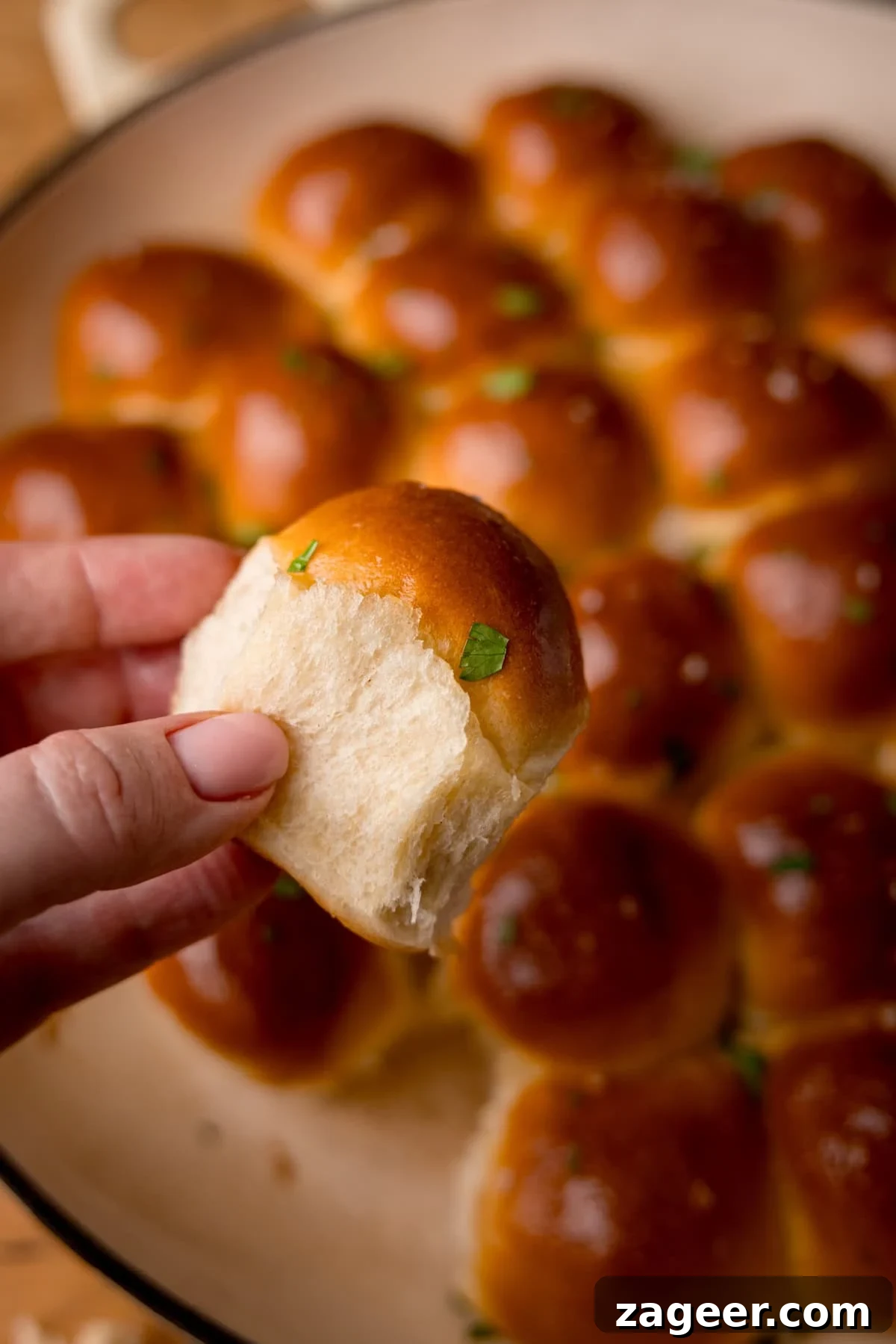 How to Make Soft, Chewy Dough Balls for Any Recipe 4 A hand holding a soft, golden-brown dough ball with a fluffy interior. In the background, a dish is filled with more dough balls, topped with chopped parsley.