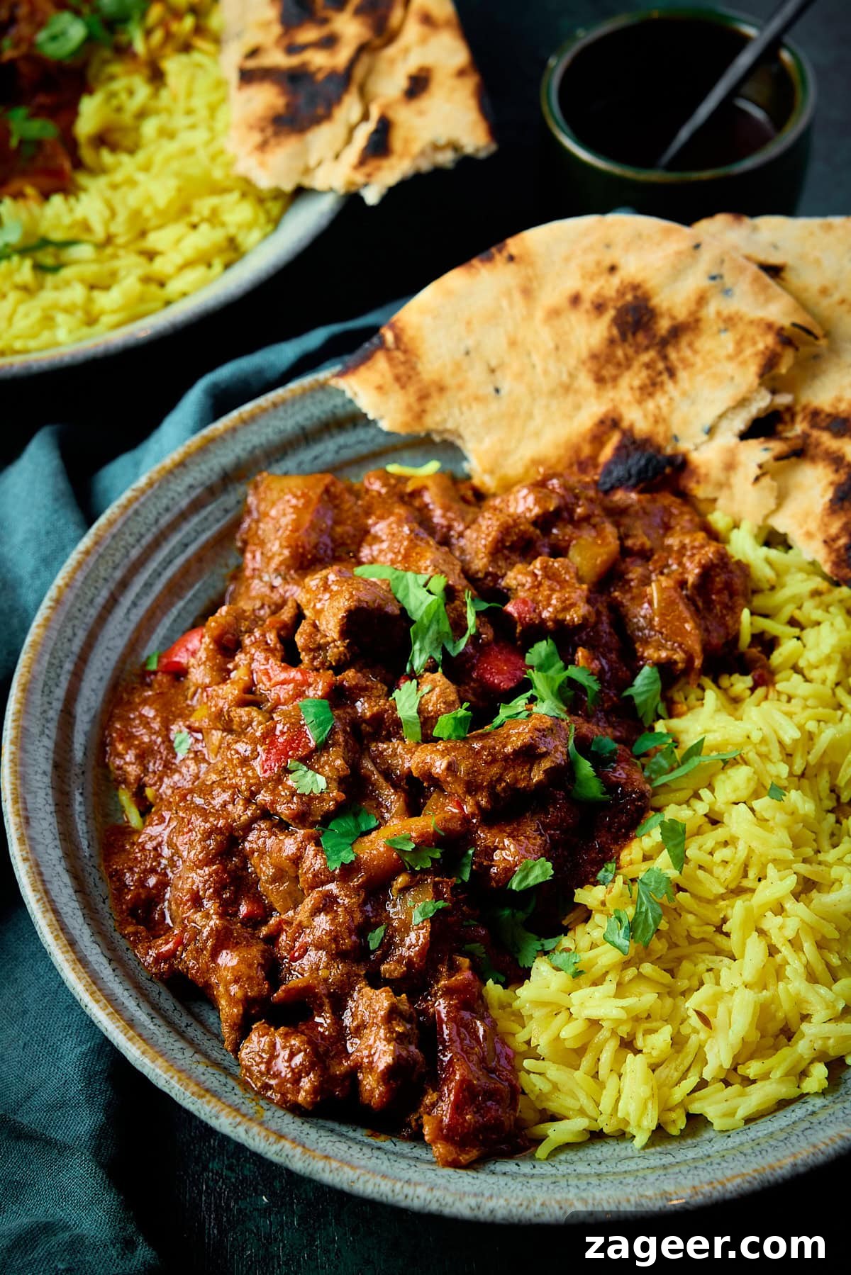 Authentic Lamb Rogan Josh Recipe: Flavorful Kashmiri Curry 4 A tall image of lamb rogan josh, in a light blue bowl with brown stripes. It is served in a bowl with pilau rice, some flatbread and topped with chopped coriander. There is also a silver spoon sticking out of the right side of the dish. In the top left corner of the image, you can see a teal napkin tucked beneath a second identical bowl of lamb rogan josh. In the right corner of the image, you can see a small black bowl with a teaspoon in it. This is all set on a teal background.