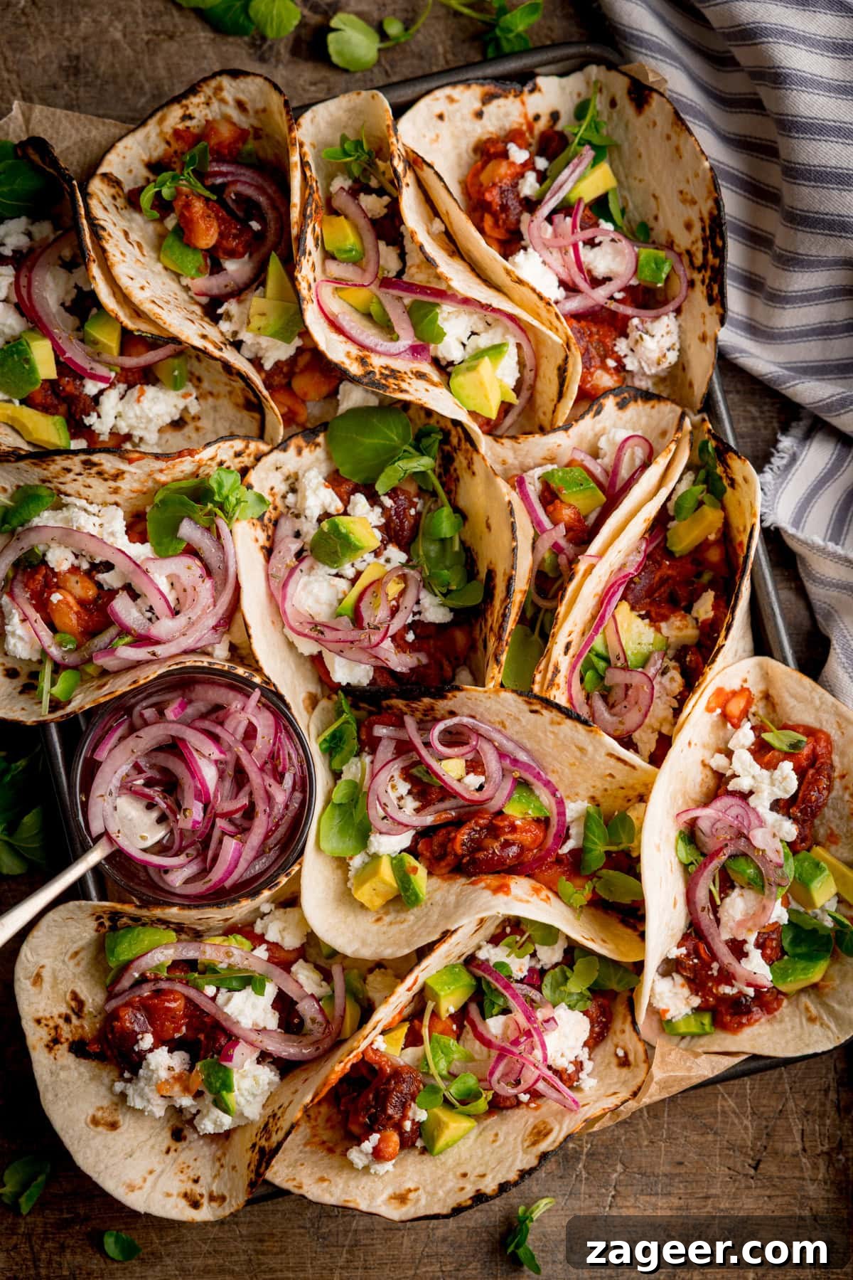 Spicy Black Bean Tacos with Fresh Salsa and Lime 2 Multiple spicy bean tacos filled with feta cheese, pickled red onions, and avocado, arranged on a tray next to a small bowl of pickled onions.
