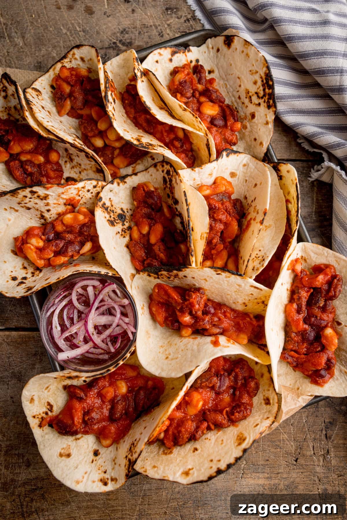 Spicy Black Bean Tacos with Fresh Salsa and Lime 6 A tray of bean tacos brimming with a savoury bean mixture, served alongside sliced red onions on a rustic wooden surface.