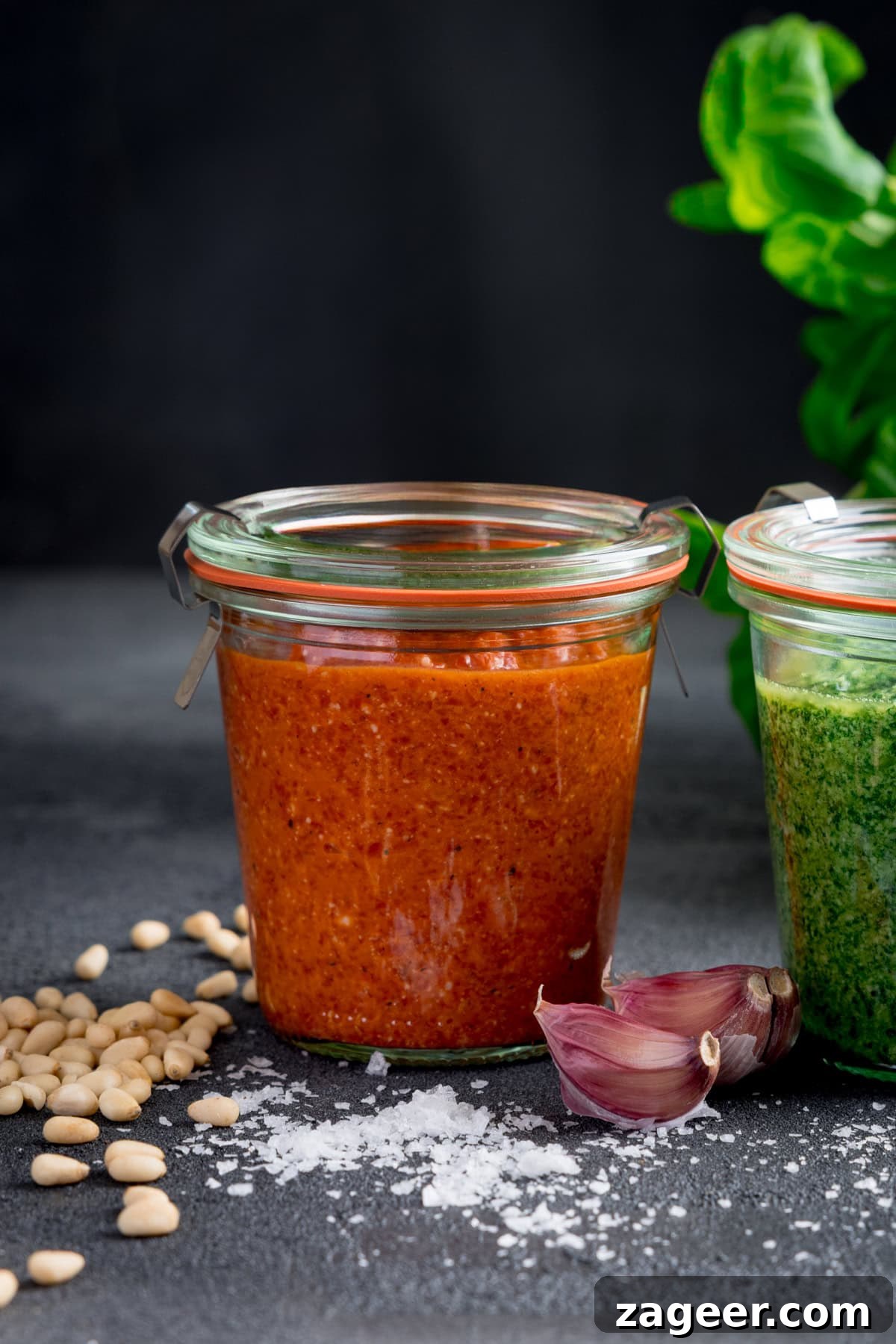 Roasted Red Pepper Pesto Recipe for Pasta, Sandwiches & Dips 2 Two glass jars of red and green pesto sit on a dark surface, surrounded by pine nuts, garlic cloves, and salt. Green leaves are visible in the background, highlighting the rich hues of the roasted red pepper pesto.