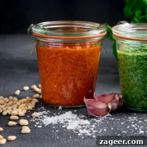 Roasted Red Pepper Pesto Recipe for Pasta, Sandwiches & Dips 6 Two glass jars, one with roasted red pepper pesto and another with green pesto, surrounded by pine nuts, garlic cloves, and coarse salt on a dark surface.