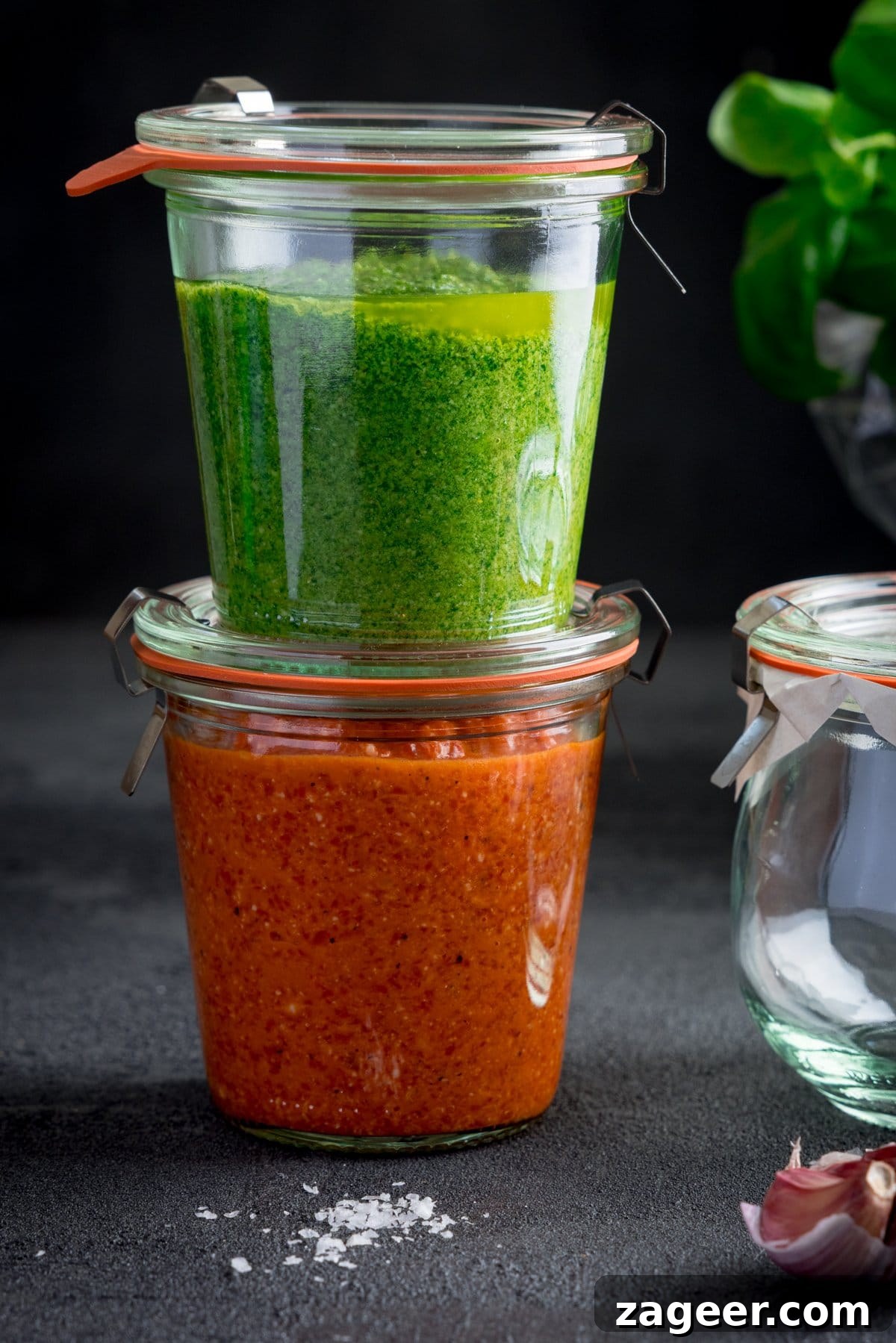 Roasted Red Pepper Pesto Recipe for Pasta, Sandwiches & Dips 4 Two sealed glass jars are stacked; the top jar reveals a vibrant green pesto while the bottom contains a red pepper pesto. The jars sit on a dark surface next to garlic cloves, a little bit of salt and an empty glass jar.