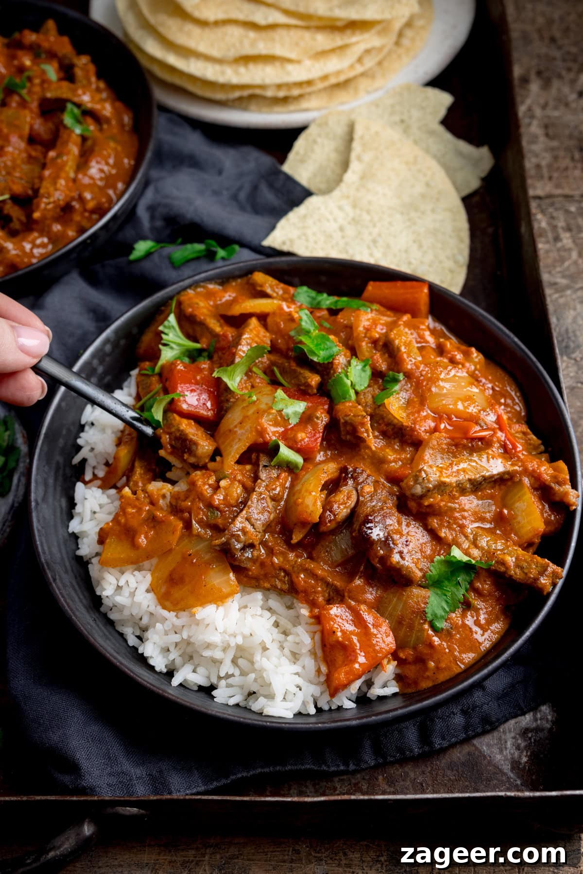 Flavorful Beef Curry Recipe: Simple Steps for Rich Homemade Curry 2 A dark bowl with boiled rice and steak curry, garnished with coriander (cilantro) on a dark tray. A forkful of the curry is being taken. There is a further bowl of curry and broken poppadoms in the background.