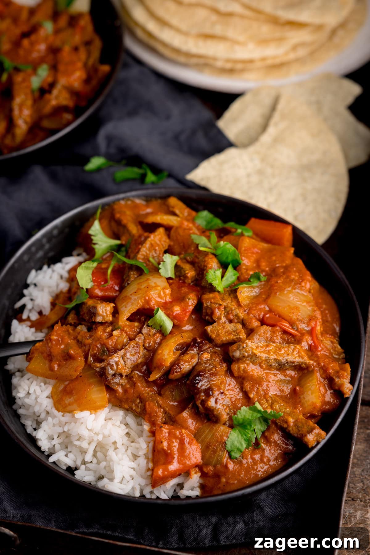 Flavorful Beef Curry Recipe: Simple Steps for Rich Homemade Curry 4 A bowl of curry and rice garnished with coriander on a dark tray. There is a plate of poppadoms and some broken up poppadoms in the background.