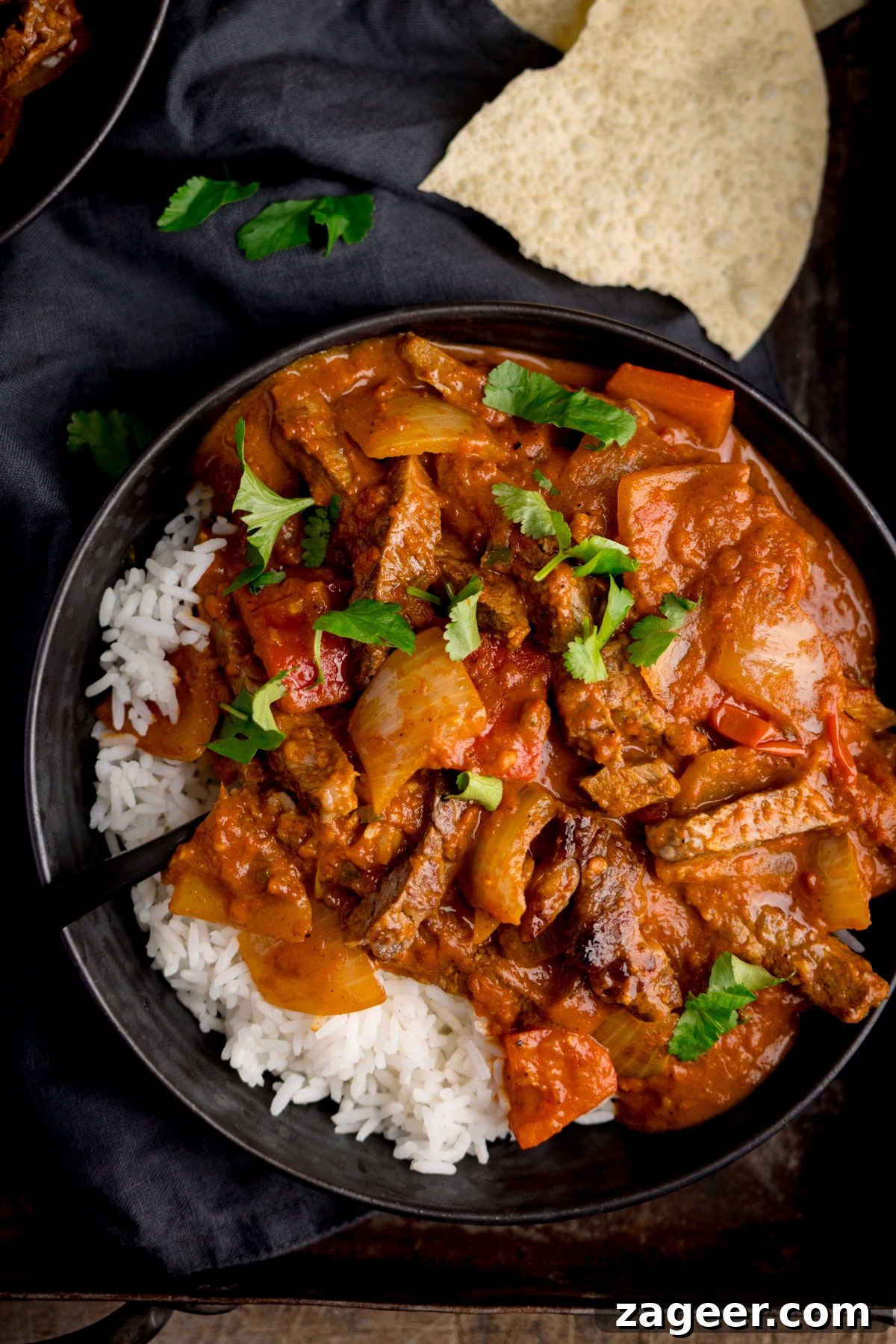 Flavorful Beef Curry Recipe: Simple Steps for Rich Homemade Curry 5 Overhead image of a bowl of curry and rice garnished with coriander on a dark tray. There are some broken up poppadoms in the background.