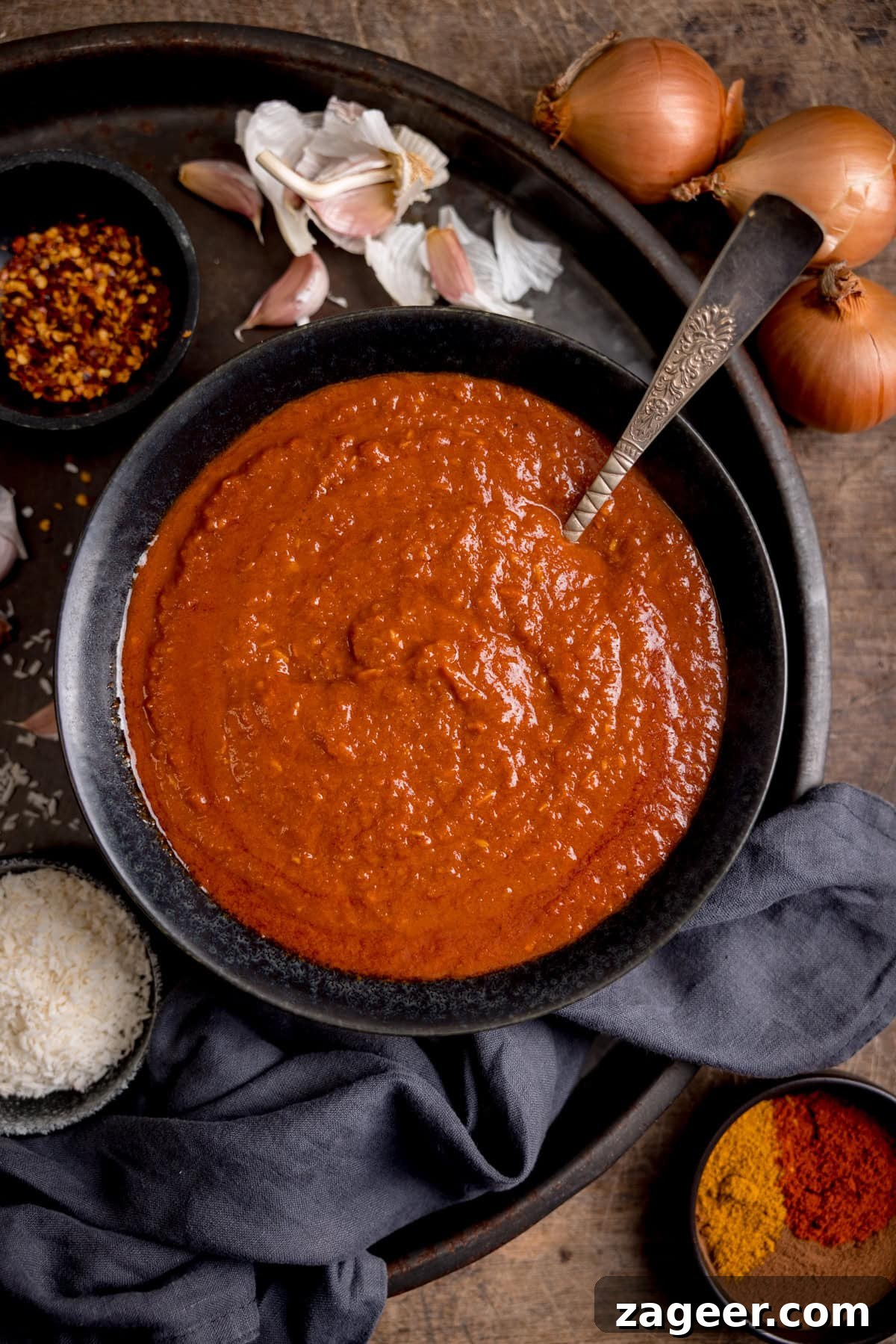 Creamy Coconut Curry Sauce Recipe for Authentic Flavor 2 A bowl of red coconut curry sauce with a spoon on a dark tray. The bowls is surrounded by spices, garlic, onions, and a grey/blue napkin.
