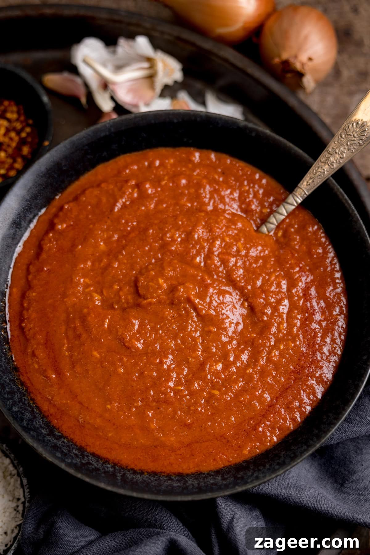 Creamy Coconut Curry Sauce Recipe for Authentic Flavor 5 A bowl of thick, red coconut curry sauce with a spoon, surrounded by garlic, onions, and a small dish of red chilli flakes.