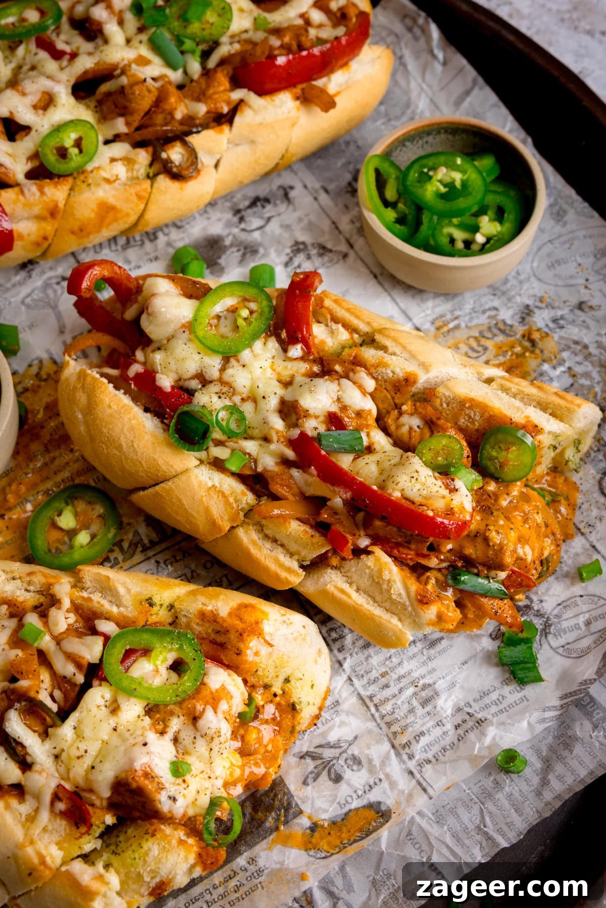 Buffalo Chicken Stuffed Garlic Bread Recipe for Game Day 4 Half of a loaded Buffalo Chicken Garlic baguette topped with melted cheese, sliced jalapeños, red peppers, and spring onions on a tray lined with printed paper; a bowl of sliced jalapeños is nearby, along with 2 further stuffed baguettes in the edges of the shot.