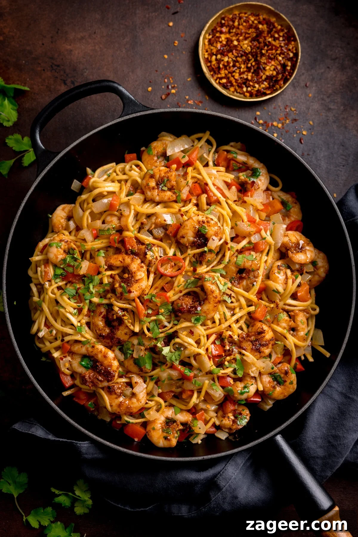Spicy Cajun Shrimp Noodles with Garlic Butter Sauce 2 A frying pan filled with Cajun King Prawns with Noodles, garnished with chopped coriander. The pan is on a dark background and there is a small bowl of crushed chilli flakes in the background.