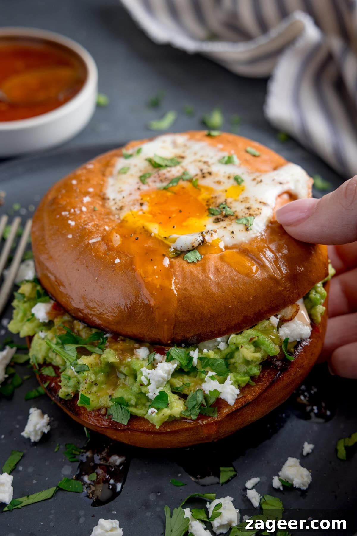 Avocado and Egg Bagel Sandwich Recipe for Breakfast 2 A hand lifts a bagel topped with a fried egg, revealing creamy avocado, feta cheese, and fresh herbs inside.