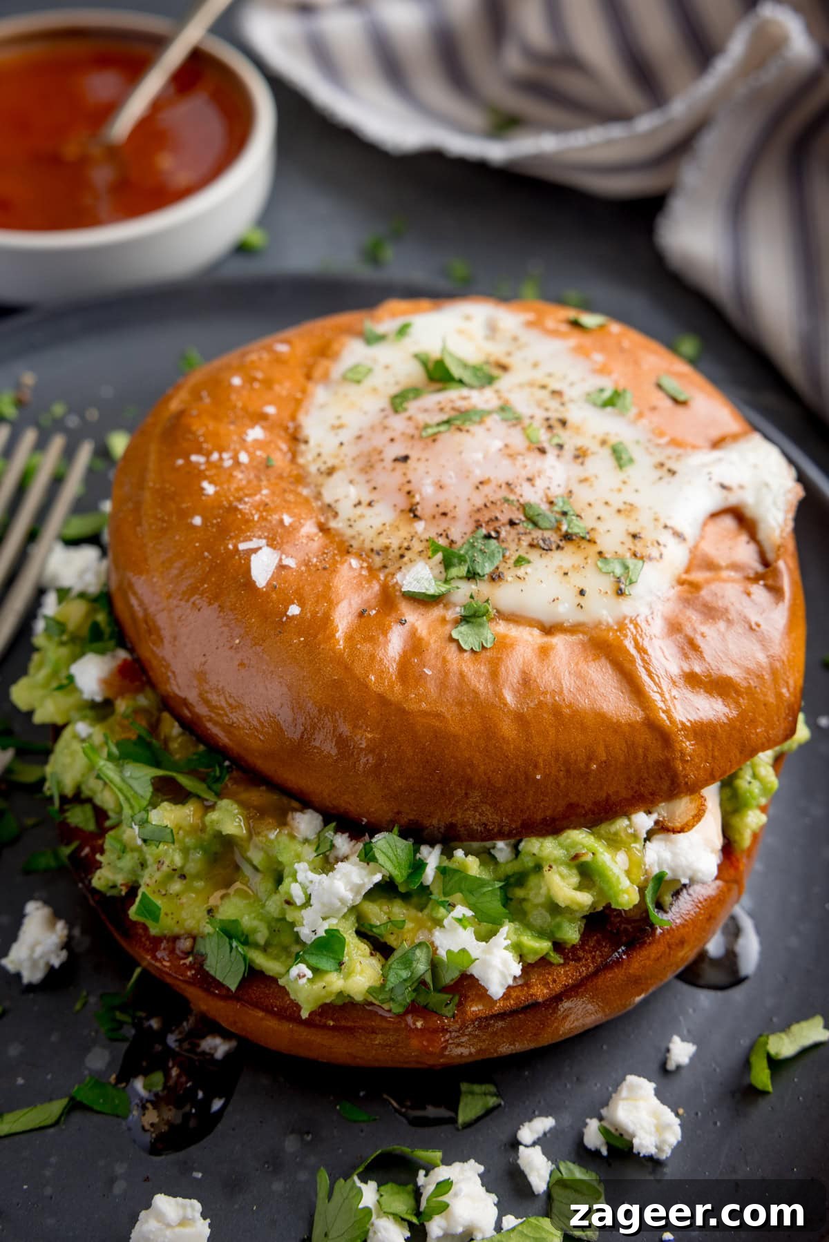 Avocado and Egg Bagel Sandwich Recipe for Breakfast 4 A bagel with a fried egg, smashed avocado, crumbled cheese, and chopped herbs sits on a dark plate with a fork beside it.