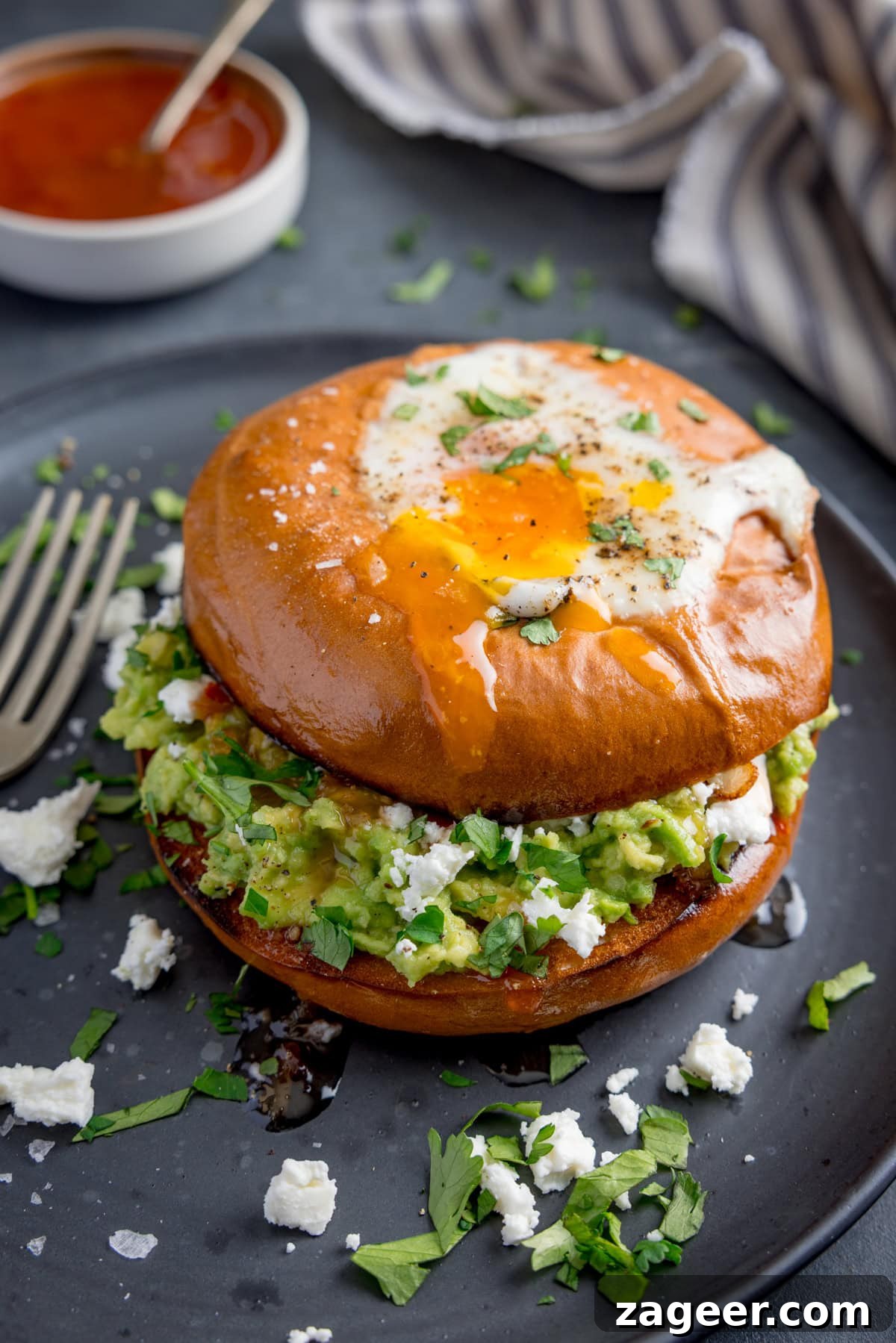 Avocado and Egg Bagel Sandwich Recipe for Breakfast 5 A bagel with a fried egg, smashed avocado, crumbled cheese, and chopped herbs sits on a dark plate with a fork beside it.