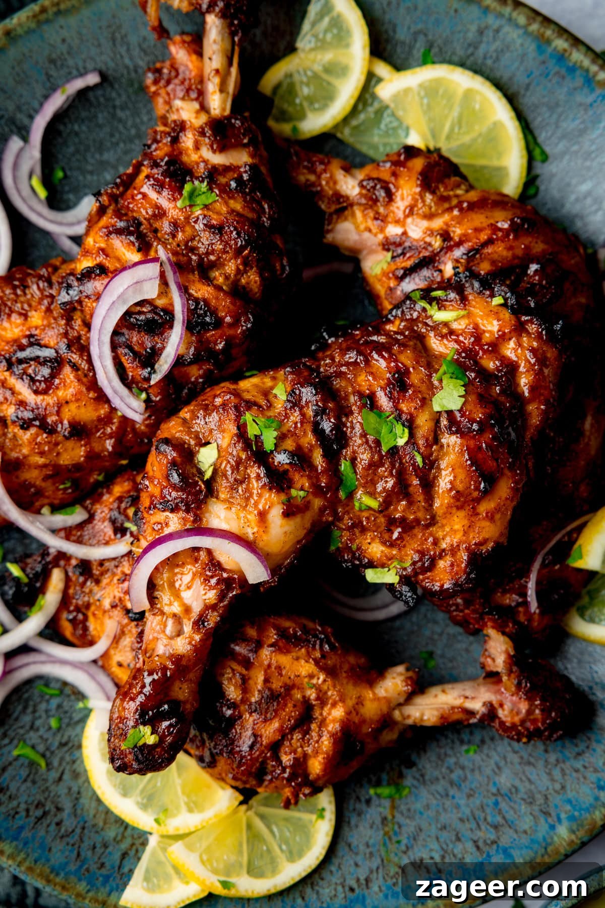 Authentic Tandoori Chicken Recipe: Flavorful Indian Grill Instructions 2 Grilled tandoori chicken legs on a blue plate. garnished with sliced red onions, fresh coriander, and lemon wedges.