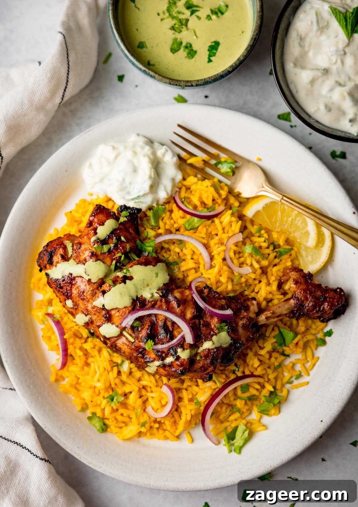 Authentic Tandoori Chicken Recipe: Flavorful Indian Grill Instructions 12 A plate of yellow rice topped with succulent tandoori chicken, red onion slices, chopped herbs, lemon wedges, and a dollop of raita, with extra sauces in bowls nearby.