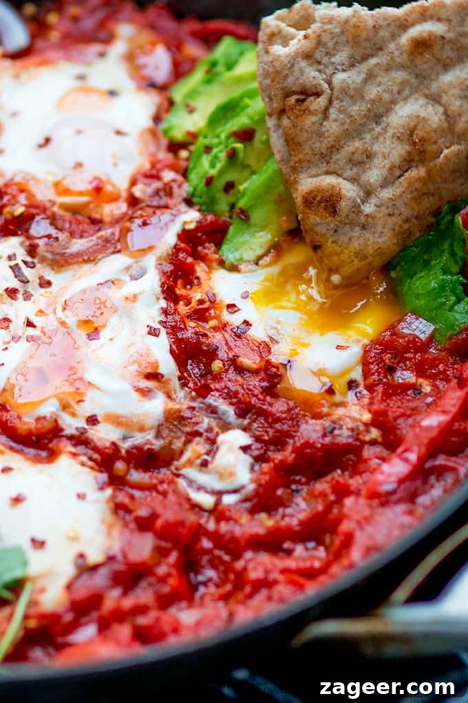 Mexican Shakshuka Recipe: Spicy Southwest Eggs in Tomato Sauce 2 Close up shot of Mexican shakshuka in a pan with avocado and chilli flakes. A piece of flatbread is being dipped in an egg yolk.