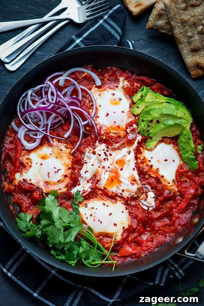Mexican Shakshuka Recipe: Spicy Southwest Eggs in Tomato Sauce 5 Overhead image of Mexican shakshuka in a pan with avocado, red onion, coriander and chilli flakes. The pan is on a dark background with flatbreads and forks just in shot.