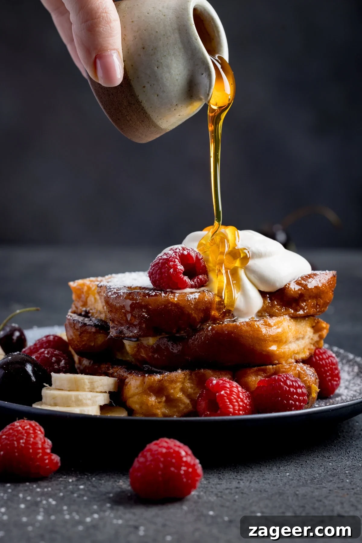 Cinnamon French Toast Recipe: Crispy, Custardy Morning Toast 2 A hand pours syrup onto a stack of caramelised French toast topped with whipped cream, raspberries, cherries, and banana slices on a black plate.