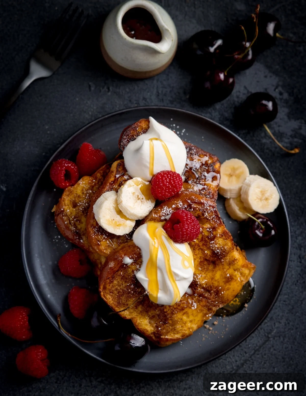 Cinnamon French Toast Recipe: Crispy, Custardy Morning Toast 5 A plate of brioche French toast topped with whipped cream, banana slices, raspberries, cherries, and syrup, with extra fruit and a small jug of syrup nearby.