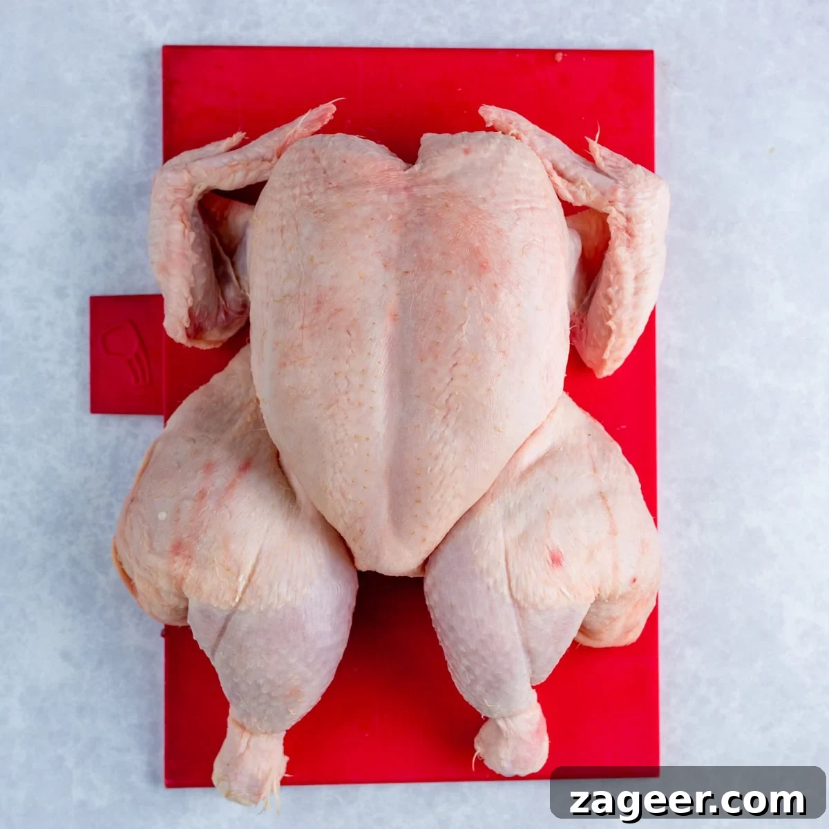 A raw whole spatchcocked chicken lying breast-side-up on a red chopping board against a light grey background.