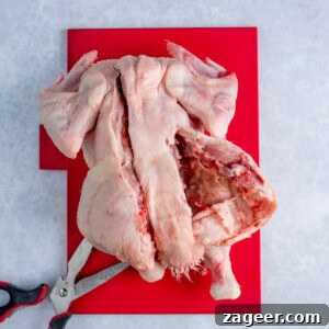 A raw butterflied chicken, partially spatchcocked with kitchen scissors, lies on a red chopping board against a light background.