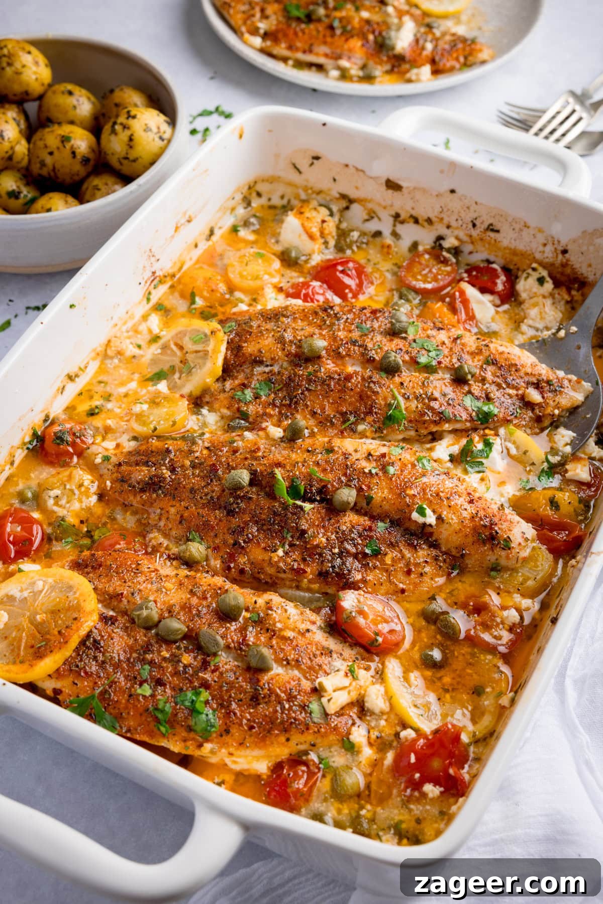 Lemon-Feta Baked Fish Recipe for Flaky, Flavorful Fillets 2 Seasoned fish fillets baked with cherry tomatoes, lemon slices, capers, and herbs in a white oven dish. A bowl of potatoes and a plate sit in the background.