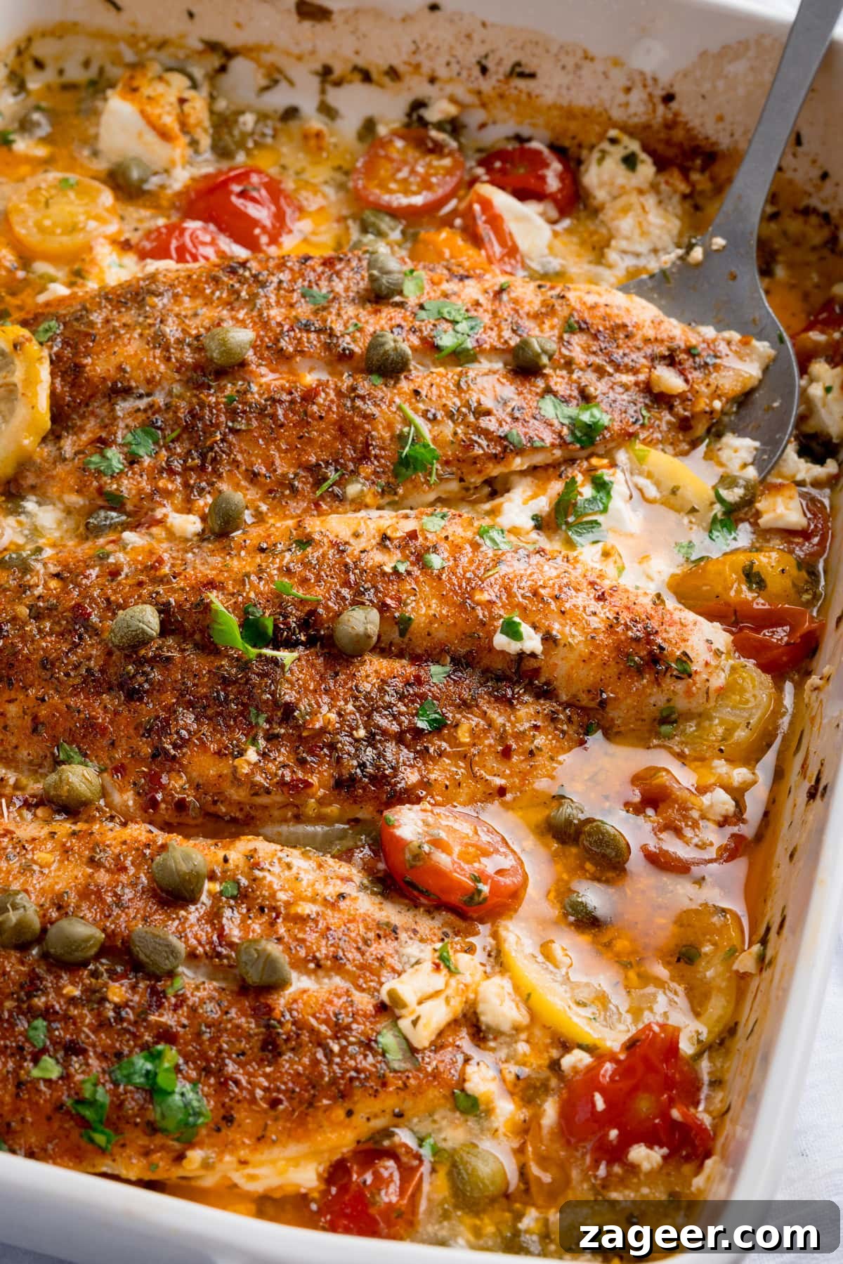 Lemon-Feta Baked Fish Recipe for Flaky, Flavorful Fillets 4 Baked Fish with Lemon and Feta fillets topped with cherry tomatoes, capers, herbs, and sauce in a white baking dish. A serving spoon is nestled in the dish.