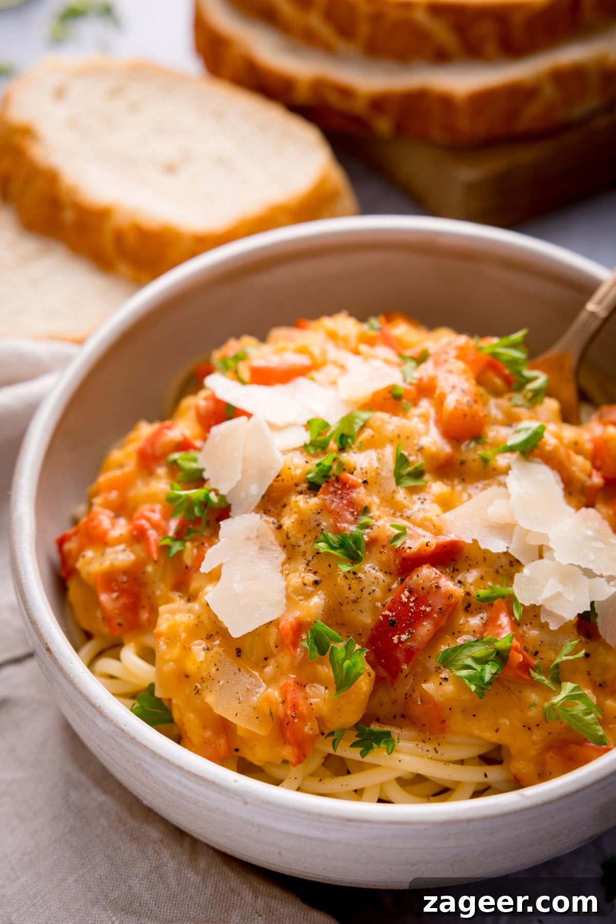 Red Pepper and Lentil Sauce Recipe for Pasta and Veggies 2 A bowl of spaghetti topped with creamy red lentil sauce, garnished with shaved parmesan and parsley, with slices of bread in the background.