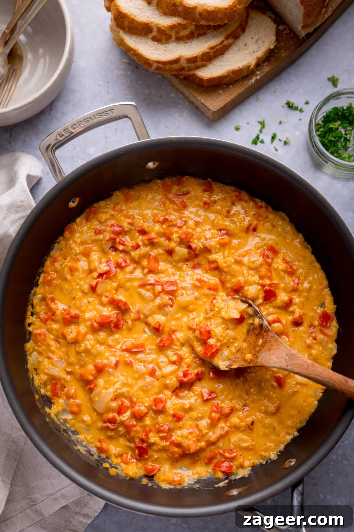 Red Pepper and Lentil Sauce Recipe for Pasta and Veggies 4 A frying pan filled with a creamy, lentil and red pepper sauce, with a wooden spoon inside. Sliced bread, herbs, and bowls are visible in the background.