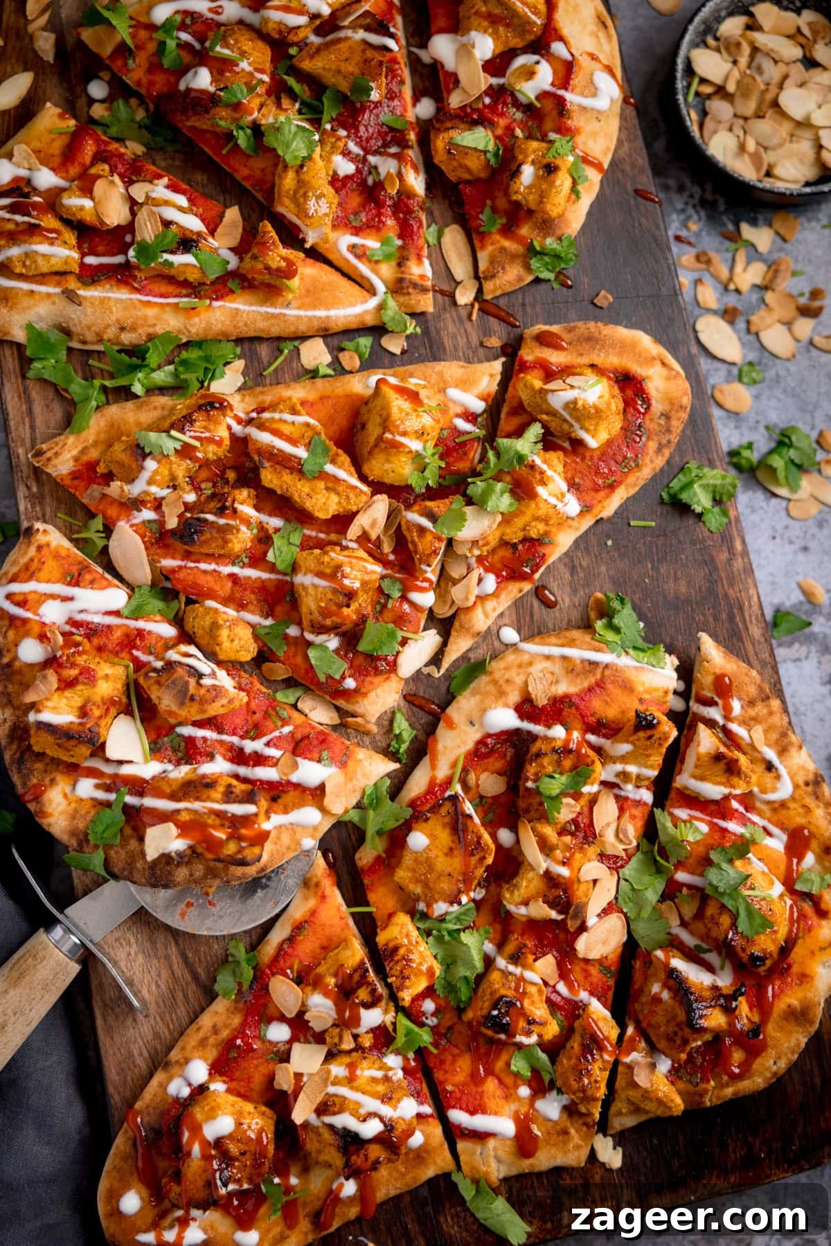Chicken Tikka Pizza Recipe: Crispy Homemade Tandoori-Style Pizza 2 Sliced chicken tikka naan pizza topped with coriander, flaked almonds, and drizzled with sauces on a wooden board. A bowl of flaked almonds sits nearby.