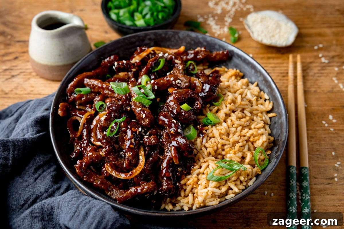 Crispy Chilli Beef in the Air Fryer: Quick Spicy Recipe 10 A wide image of a black bowl filled with air fryer crispy chilli beef with onions, sesame seeds, and sliced spring onions, served with fried rice. The bowl is on a wooden surface next to a blue/grey napkin, a pair of chopsticks, a small jug of soy sauce, a small bowl of chopped spring onions and a small dish of sesame seeds.