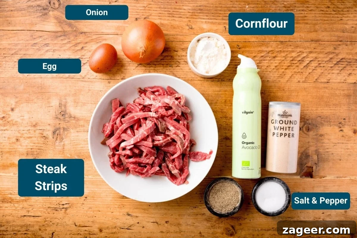 Crispy Chilli Beef in the Air Fryer: Quick Spicy Recipe 4 Ingredients for Air Fryer Crispy Chilli Beef on a wooden surface. The ingredients are labelled, including: a bowl of raw steak strips, egg, onion, cornflour, salt, pepper, white pepper and avocado oil spray
