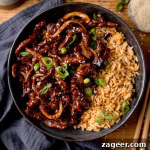 Crispy Chilli Beef in the Air Fryer: Quick Spicy Recipe 7 A black bowl filled with air fryer crispy chilli beef with onions, sesame seeds, and sliced spring onions, served with fried rice. The bowl is on a wooden surface next to a blue/grey napkin, a pair of chopsticks and a small dish of sesame seeds.