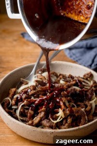 Crispy Chilli Beef in the Air Fryer: Quick Spicy Recipe 9 Chilli beef sauce is being poured over a bowl of crispy chilli beef strips and sliced onions on a wooden table.