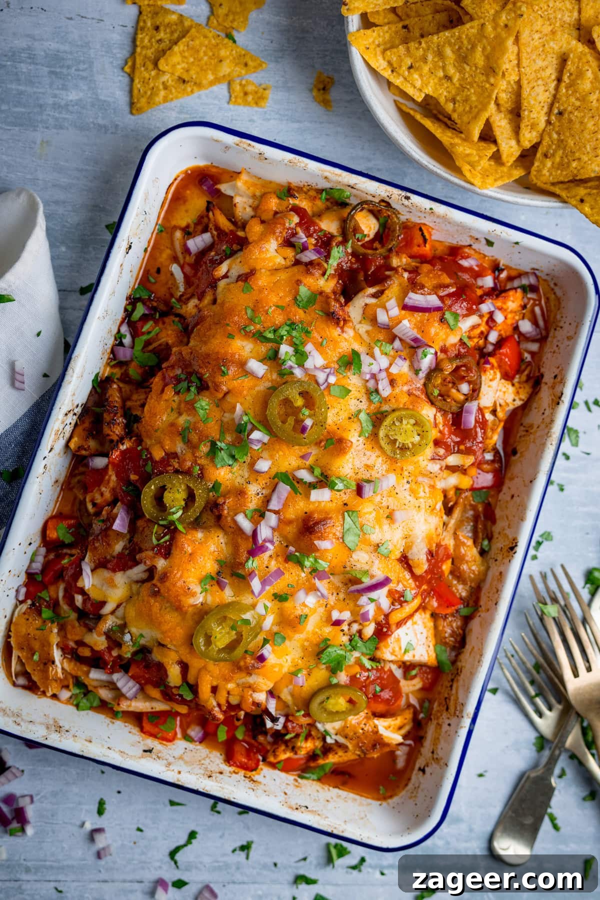 Slow-Cooker Salsa Chicken Recipe for Flavor-Packed Weeknight Meals 2 A baking dish filled with cheesy chicken salsa bake, topped with jalapeños, red onions, and chopped herbs, with a bowl of tortilla chips and cutlery nearby on a light surface.