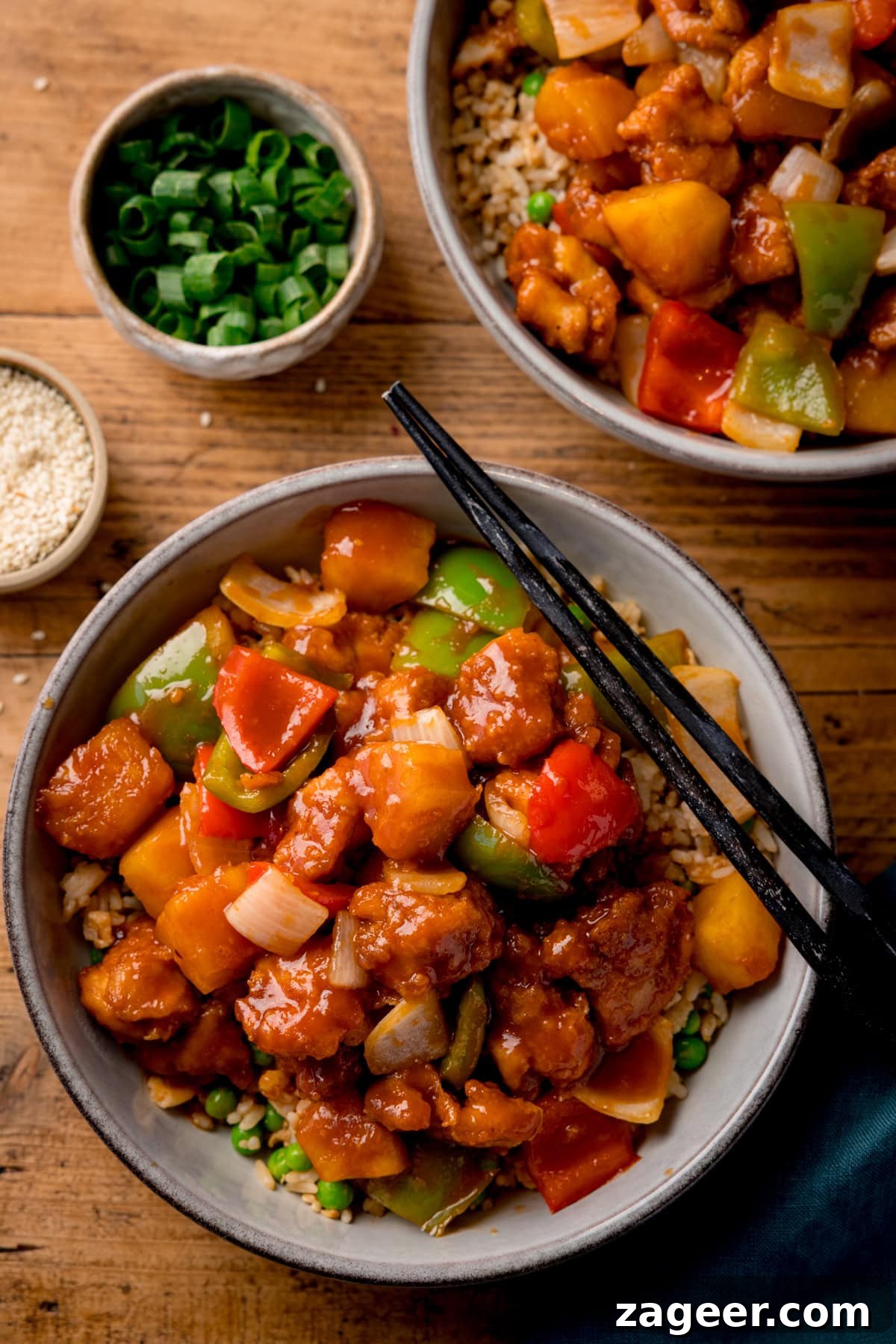 Crispy Air Fryer Sweet and Sour Chicken Recipe 2 Tall overhead image of a bowl of sweet and sour chicken with chunks of onion, red & green peppers and pineapple in sauce. The bowl is on a wooden surface and a pair of black chopsticks rest on the bowl. A further bowl of sweet and sour chicken is partly in shot at the top of the frame. Small bowls of chopped green onion and sesame seeds are nearby.