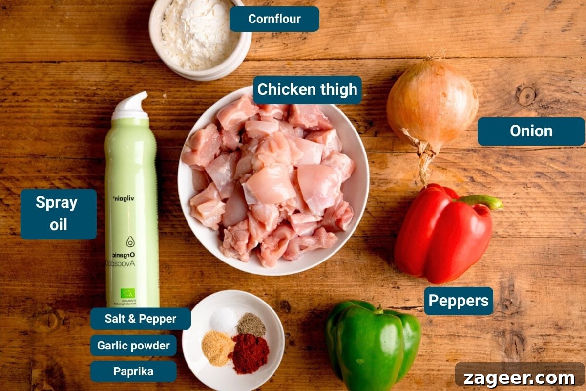 Crispy Air Fryer Sweet and Sour Chicken Recipe 4 Chopped chicken thigh in a bowl, cornflour, spray oil, onion, red and green peppers, and spices on a wooden table. Each item is labelled.
