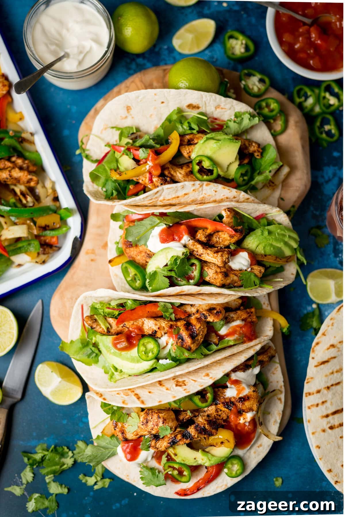 Quick Chicken Fajitas with Peppers and Onions 2 Three grilled chicken fajitas with peppers, onions, avocado, jalapeños, salsa and soured cream on tortillas, arranged on a blue surface with limes, salsa, and coriander nearby.