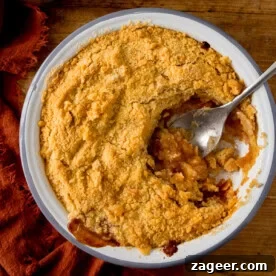 15 Cozy Autumn Baking Ideas for Weekend Treats 4 A round baking dish filled with golden-brown apple crumble. A scoop is being taken by a silver serving spoon. The dish is on a wooden surface next to a terracotta napkin.