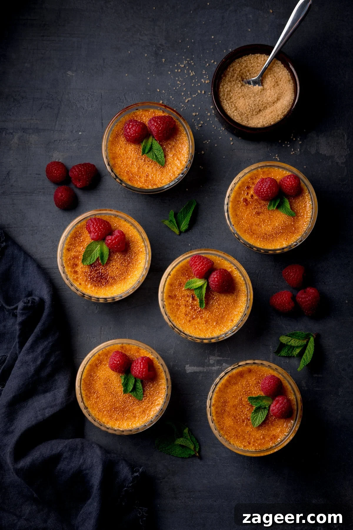 Classic Crème Brûlée Recipe: Silky Custard with Caramelized Sugar 2 Six ramekins of crème brûlée topped with raspberries and mint, surrounded by loose raspberries, mint leaves, a bowl of brown sugar, and a dark cloth on a dark surface.