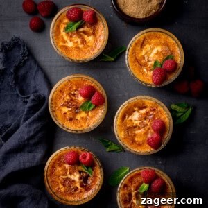 Classic Crème Brûlée Recipe: Silky Custard with Caramelized Sugar 11 Six ramekins of crème brûlée topped with fresh raspberries and mint leaves are arranged on a dark surface, with a bowl of brown sugar and a dark serviette nearby.
