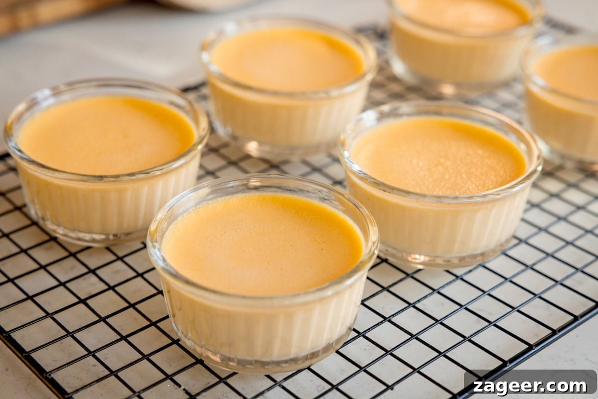 Classic Crème Brûlée Recipe: Silky Custard with Caramelized Sugar 6 Six glass ramekins filled with smooth, yellow custard are cooling on a black wire rack set on a light-coloured worktop.