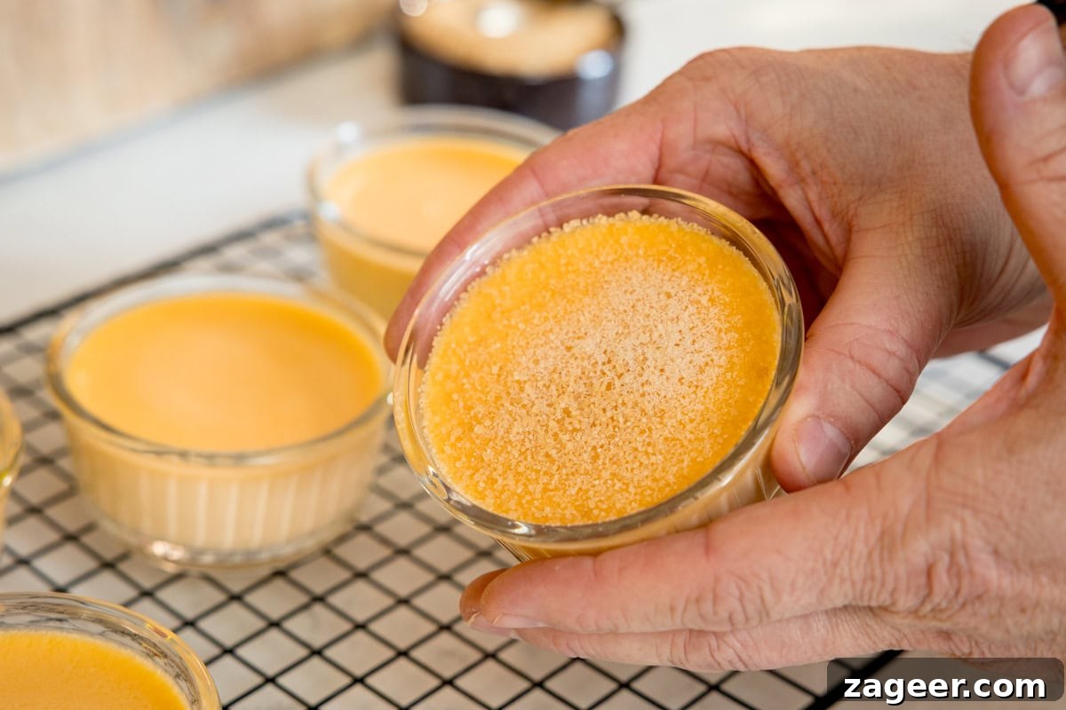 Classic Crème Brûlée Recipe: Silky Custard with Caramelized Sugar 7 A person holds a small glass dish of custard with sugar sprinkled on top, with more custard dishes cooling on a wire rack in the background.