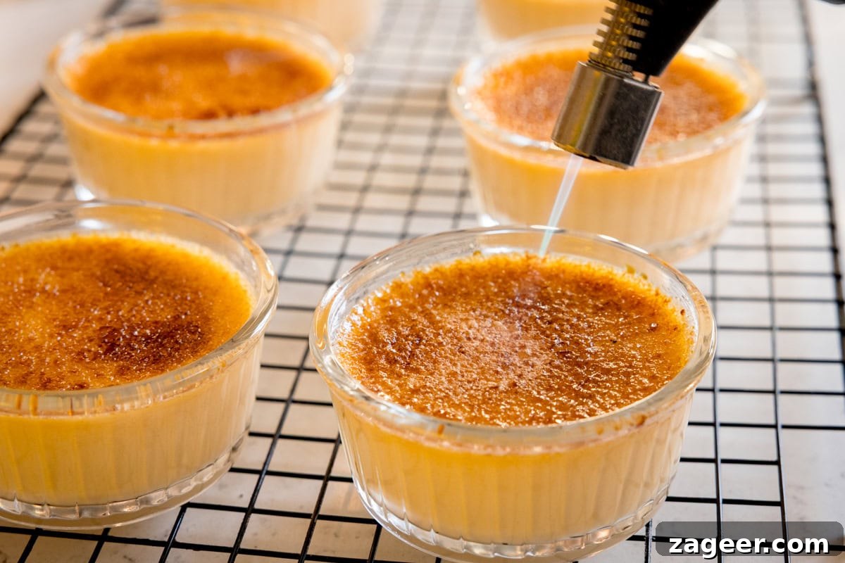 Classic Crème Brûlée Recipe: Silky Custard with Caramelized Sugar 8 Several ramekins of crème brûlée are shown, with a kitchen blowtorch caramelising the sugar on top of one, all on a cooling rack.