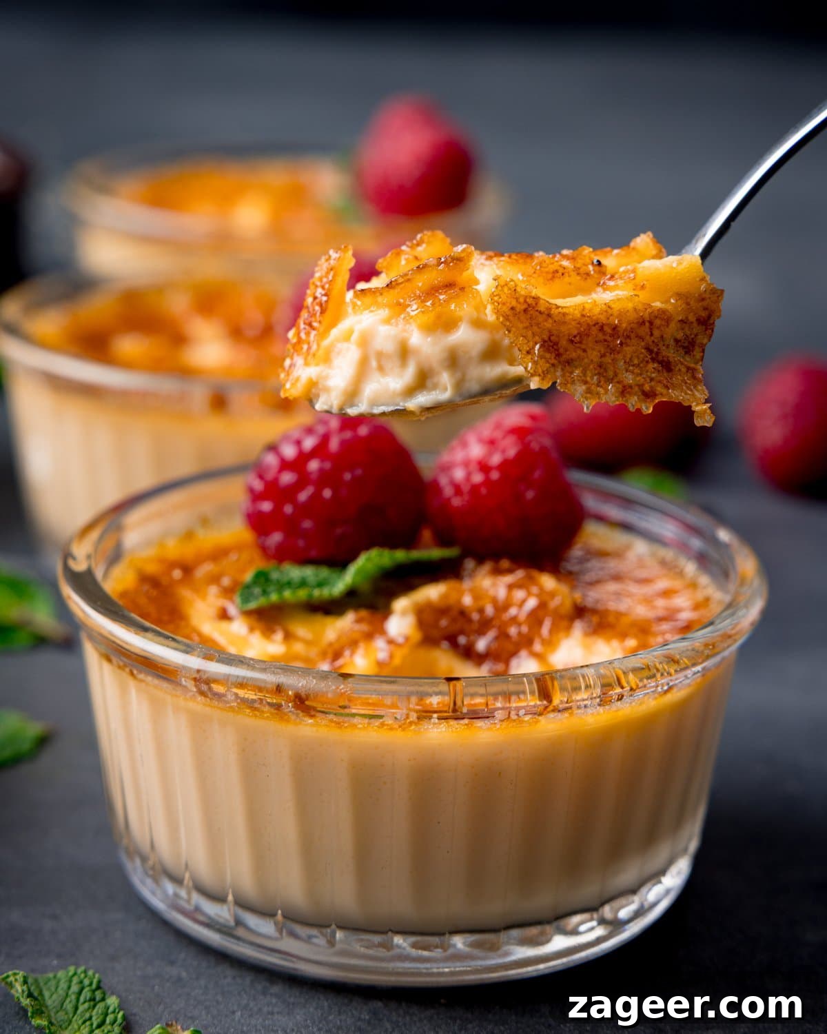 Classic Crème Brûlée Recipe: Silky Custard with Caramelized Sugar 10 A spoon lifts a bite of crème brûlée from a glass dish, topped with caramelised sugar, fresh raspberries, and mint. Two more servings appear in the background.