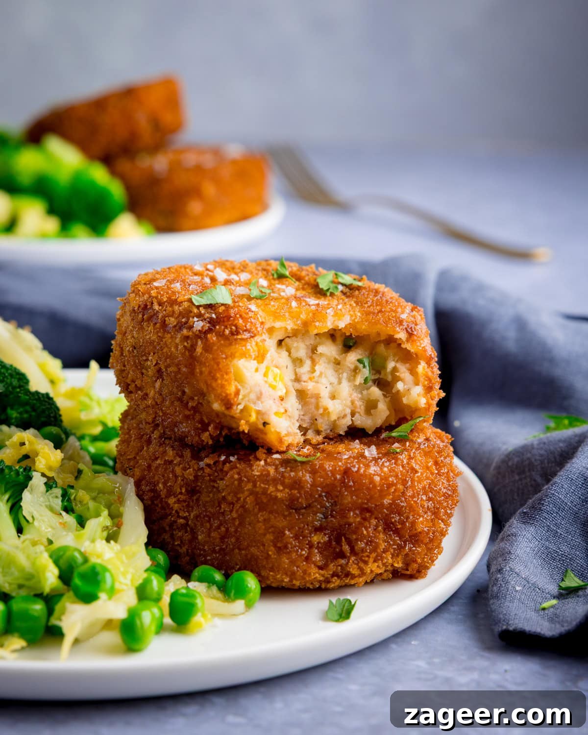 Crispy Tuna Fish Cakes with Lemon-Dill Sauce 7 Two breaded tuna fish cakes are stacked on a plate, one partially eaten, served with peas, broccoli, and shredded cabbage.