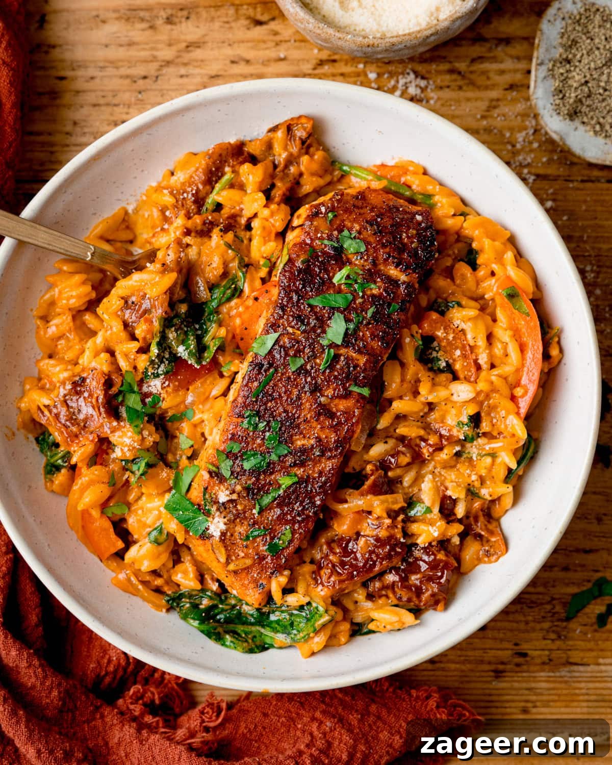 Tuscan Salmon with Creamy Lemon Orzo Recipe 2 Tall overhead image of a white bowl with Tuscan-style orzo and pan fried salmon on top, garnished with parsley. The bowl is on a wooden table next to an orange napkin.
