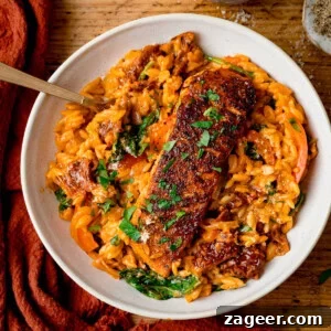 Tuscan Salmon with Creamy Lemon Orzo Recipe 7 Square image of a white bowl with Tuscan-style orzo and pan fried salmon on top, garnished with parsley. The bowl is on a wooden table next to an orange napkin.